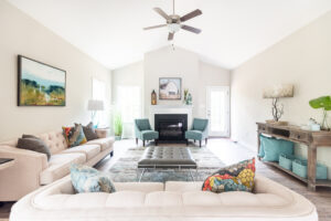 Bright, spacious living room with a beige sofa, colorful cushions, a central ottoman, a fireplace, and tasteful decor, illuminated by natural light.