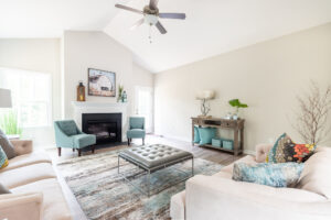 Bright, contemporary living room with teal armchairs, a gray sofa, ottoman, fireplace, and a glass-top coffee table.