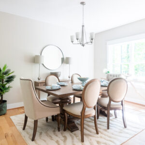 Elegant dining room with a wooden table set for four, surrounded by upholstered chairs, under a modern chandelier, adjacent to a bright window and a circular mirror on the wall.