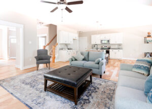 Bright, spacious living room with a connected kitchen, featuring white cabinetry, hardwood floors, and modern furnishings including sofas and a leather ottoman.
