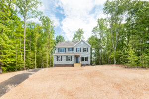 A two-story, gray house with white trim is surrounded by trees and sits on a newly landscaped yard with a paved driveway.