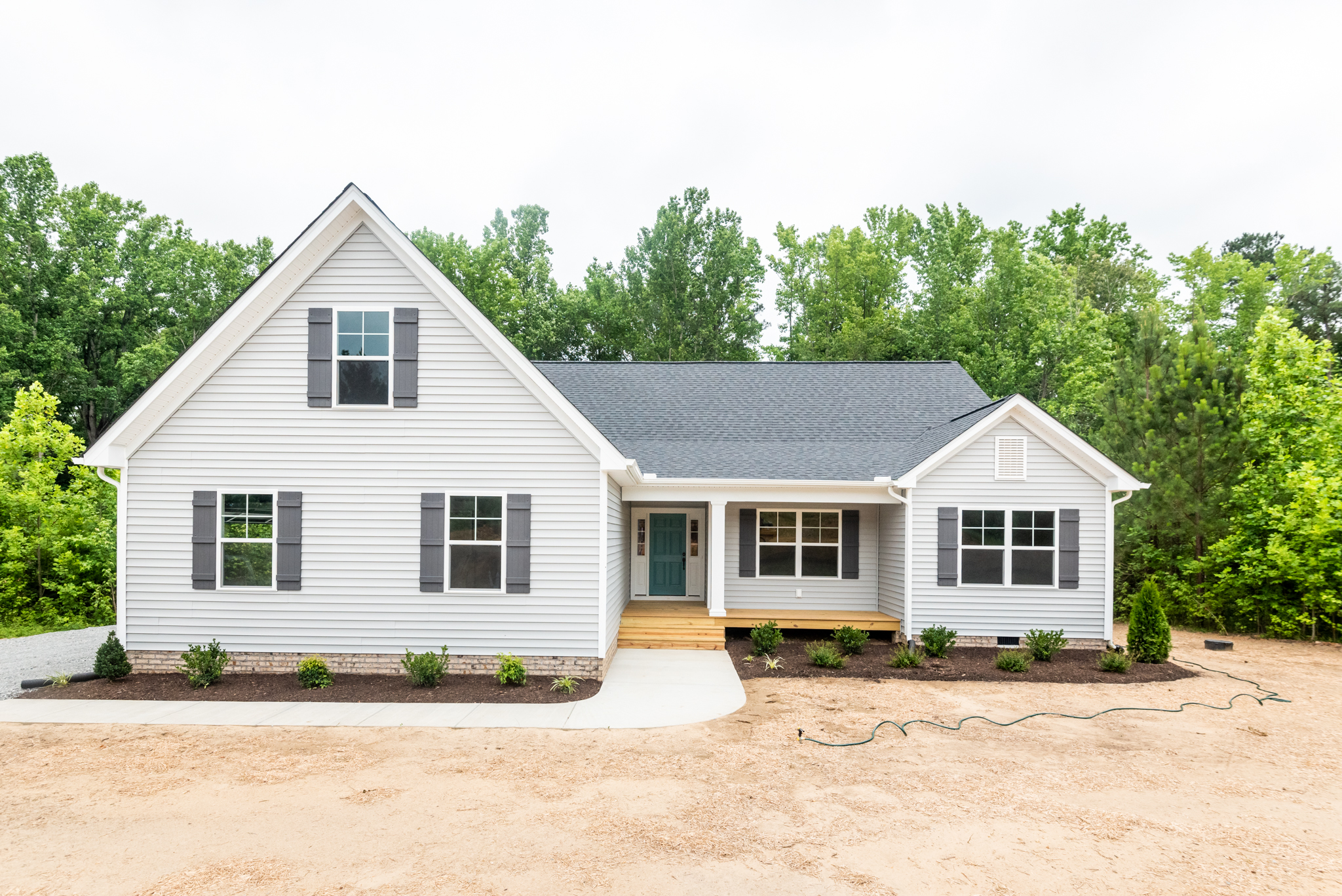 A new single-story house with white siding, gray roof, and a covered front entrance, surrounded by a freshly landscaped yard and a backdrop of trees.