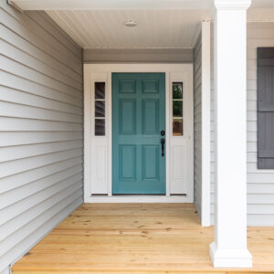 A teal front door of a house with light gray siding, flanked by white columns and a wooden floor porch.