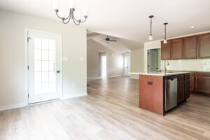 Spacious kitchen area with wooden floors, brown cabinets, an island with a sink, and a light fixture. In the background, there is a bright, empty room and a door leading outside.
