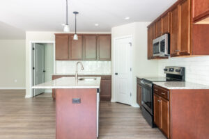 A modern kitchen featuring wooden cabinets, stainless steel appliances, a center island with a sink, pendant lighting, a white subway tile backsplash, and hardwood floors.