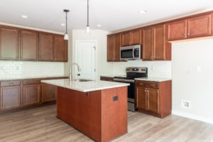 A modern kitchen with brown cabinets, a center island with a sink, white tile backsplash, stainless steel appliances, and wooden flooring.
