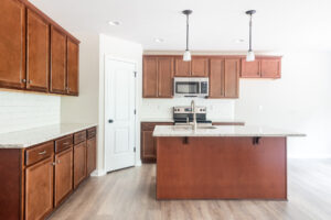 A modern kitchen with wooden cabinets, a center island, stainless steel appliances, and pendant lights. The space features light-colored countertops and a white door on the left.