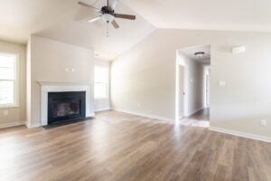 A bright, empty room with light wood flooring, a vaulted ceiling with a ceiling fan, a fireplace, and a hallway leading to other areas. The walls are neutral-colored and two windows allow natural light.