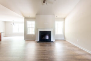 A spacious empty room with light wood flooring, neutral wall colors, and a central fireplace. Large windows on either side of the fireplace allow natural light to fill the space.