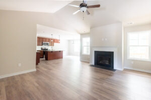 A spacious, empty living area with wood flooring, a ceiling fan, recessed lights, a fireplace, and large windows. The kitchen with cabinets and a counter is visible in the background.