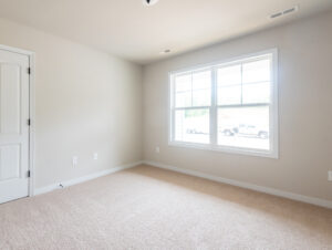 Empty room with beige walls and carpet, a large window letting in natural light, and a white door on the left wall.