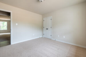 Empty beige-walled room with a closed white closet door, beige carpet, and adjacent room visible through an open entryway.