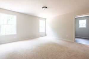 An empty, well-lit room with beige carpet, white walls, two windows, and a ceiling light fixture. A doorway on the right leads to another room.