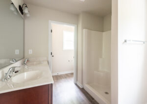 A clean bathroom with a double sink vanity, a step-in shower, a towel bar on the wall, and an open doorway leading to a room with a window.