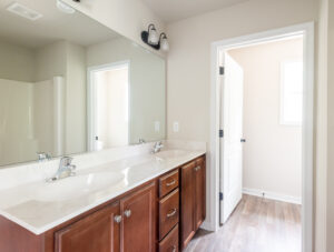 A bathroom with a double sink vanity, large mirror, and wooden cabinets. The room features a doorway leading to another area, light-colored walls, and wood-look flooring.