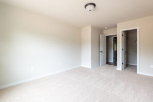 A vacant, carpeted room with plain white walls, a ceiling light, and two open doors leading to a closet and a hallway.