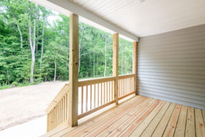 A wooden deck with a railing and stairs leading to an open area, adjacent to a forested backdrop. The deck is attached to a house with light gray siding and is sheltered by a white ceiling.