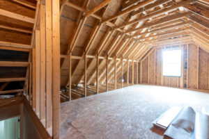 A spacious, unfinished attic with exposed wooden beams, plywood flooring, and a window providing natural light. Rolls of insulation are placed on the floor.