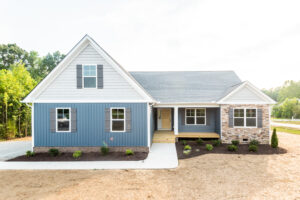 A new single-story blue house with gray shingles, white trim, stone accents, a driveway, and freshly landscaped garden.