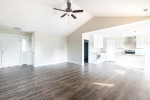 Spacious, empty room featuring dark wood flooring, white walls, and a modern kitchen with white cabinets in the background. a ceiling fan hangs overhead.