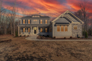 A two-story house with front porch and multiple windows is set against a vibrant sunset sky. A curved pathway leads to the entrance, and the yard is devoid of plants. Trees surround the property.