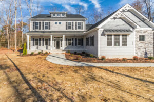 A newly constructed two-story house with light grey siding, dark roof, and white trim. The house features a front porch, multiple windows, and a landscaped yard on a clear day.
