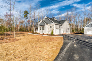 A newly constructed two-story house with a gray exterior and stone accents, set against a backdrop of leafless trees. The property includes a curved driveway leading to a garage.