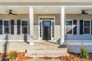 A front porch of a gray house with white pillars, a black front door, and two ceiling fans. The steps are brick, and dried leaves are scattered on the ground.