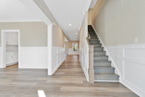 A hallway with light wood flooring and white trim features a staircase on the right and leads to a kitchen area on the left. The space is bright and modern with neutral-colored walls.
