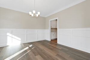 A bright dining room with beige walls, white wainscoting, a hanging light fixture, and wood flooring. An open doorway leads to an adjacent room with white cabinetry.