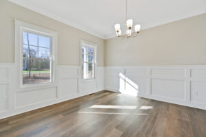 A bright, empty room with beige walls, white wainscoting, hardwood floor, three windows, and a chandelier.
