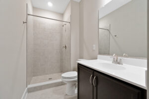 A modern bathroom with a glass-front shower, toilet, and dark wood vanity with a white countertop and sink, located next to a large mirror. The wall and floor tiles are beige.