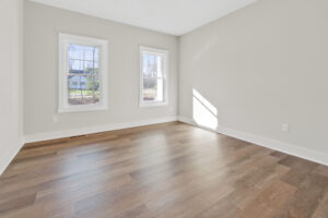 An empty room with pale walls, two windows, and wood flooring. Natural light is entering from the windows, creating bright spots on the floor.