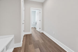 Empty hallway with light-colored walls and wood flooring, leading to a bathroom visible through an open door.