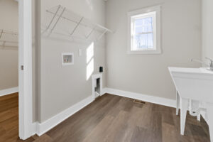 A laundry room with wood flooring, wall-mounted wire shelving, a window, utility sink, and plumbing connections for washer and dryer.