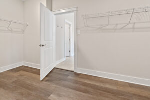 Empty walk-in closet with open door leading to a lit hallway, featuring wooden flooring and metal wire shelving on beige walls.