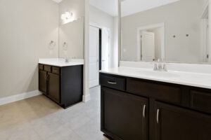 A bathroom with two dark wood vanities, each with a sink and a mirror, light gray tiled flooring, and light beige walls.