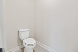 A clean, white toilet is positioned against the light gray wall in a small, sparsely decorated bathroom.