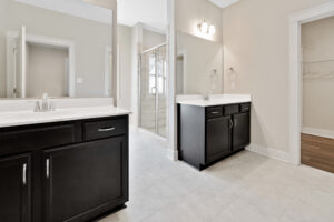 Modern bathroom with twin dark wood vanities, white countertops, a glass-enclosed shower, light-colored tile flooring, and beige walls.