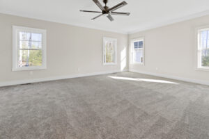 A spacious room with beige walls, three windows, gray carpet, and a ceiling fan. Natural light enters through the windows.