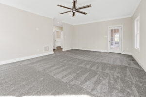 A spacious, empty room with light gray carpet, white walls, a ceiling fan, and a door leading to another room.