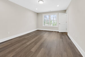 An empty room with light gray walls, a wooden floor, a central ceiling light, a single closed white door, and a large window showing trees outside.