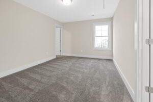 A vacant room with beige walls, white trim, a window, and gray carpet. A closed door is visible on one side.