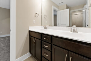 A bathroom with a double sink vanity, dark wood cabinets, and silver fixtures. A partially open door reveals a glimpse of a shower and adjacent carpeted room.