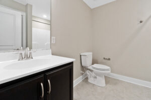 Modern bathroom with a sink on the left, a toilet on the right, and beige walls. The floor is tiled, and there is a towel hook on the wall.