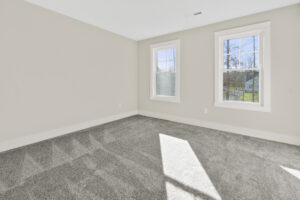 An empty room with light gray walls and carpet, and two windows allowing natural light.