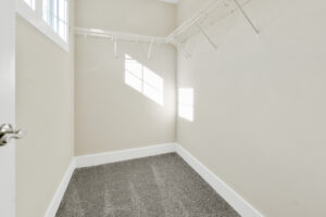 Empty walk-in closet with beige walls, gray carpet, and white wire shelving. Sunlight enters through a window, casting grid-like shadows on the wall.
