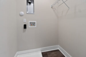 Empty laundry room with white walls, a wall-mounted shelf, outlets, and plumbing fixtures. Dark flooring is visible at the bottom.