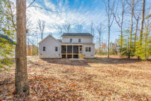 A two-story house with a porch is surrounded by trees and fallen leaves under a blue sky.