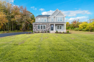 A modern two-story house with gray siding and a manicured lawn, situated next to a wooded area under a blue sky.